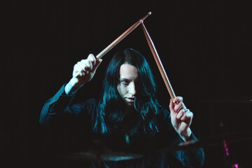 A female metal drummer with long hair holding up drumsticks behind the drumkit against a dark background - obrazy, fototapety, plakaty