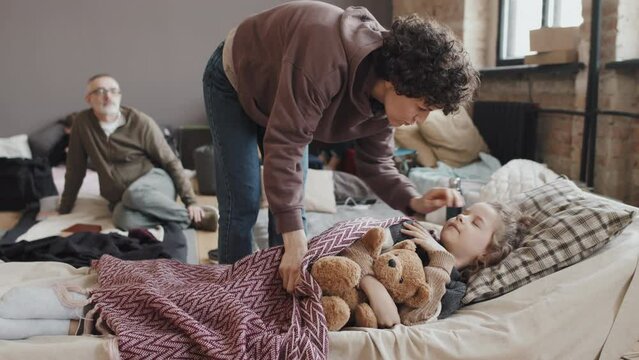 Wide shot of curly young Caucasian woman putting blanket on child and kissing him on forehead, boy napping on cot in refuge at daytime