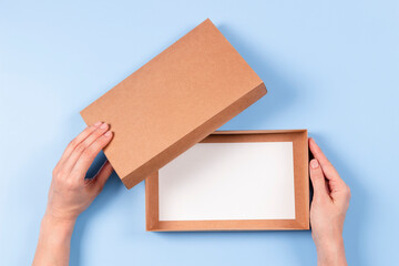 Top view to female hands open empty brown cardboard box on light blue background. Mockup parcel...