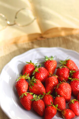 Lilac plate full of fresh strawberries, open book and reading glasses on wooden table. Selective focus.