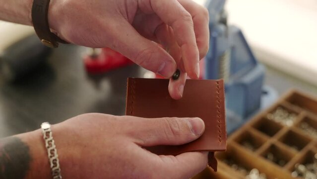 Leather craftsman works in workshop. Master making leather wallet. Man installs tack button snap fastener fitting using hand press grommet machine. Male using manual punch tool for fastener sewing.