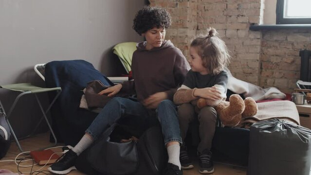 Full shot of curly Caucasian woman sitting on cot in shelter at daytime, unpacking her bag, cute little boy with stuffed bear hugging mother