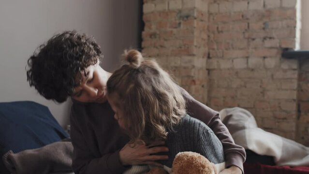 Tracking low angle of cute Caucasian little boy carrying stuffed bear, walking in indoor shelter at daytime, snuggling with curly-haired young mother