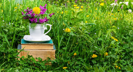 a white teacup with a bouquet of wildflowers on a stack of old books in a meadow with green grass on a sunny spring or summer morning