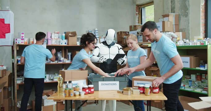 Automatic robot making notes on tablet while group of people packing donation boxes indoors. Team of young volunteers with technological cyborg working at food bank warehouse.