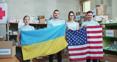 Team of food bank workers in volunteer wear holding Ukraine and USA flags while standing at warehouse of charity foundation. American volunteers preparing humanitarian aid for delivering in Ukraine.