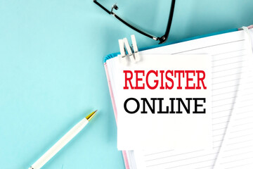 REGISTER ONLINE text on a sticky on notebook with pen and glasses , blue background