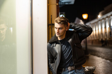 Fototapeta Fashionable handsome young man model with hairstyle in a stylish black leather jacket walking in a night city and standing near a shop window obraz