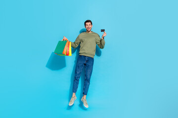 Full size photo of impressed young brunet guy jump with bags card wear shirt jeans shoes isolated on blue background