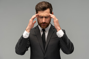 Unhappy frustrated bearded man wearing official style suit suffering migraine or high blood pressure while massaging temples and feeling headache. Indoor studio shot isolated on grey background
