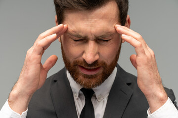 Unhappy frustrated bearded man wearing official style suit massaging temples feeling headache, suffering migraine or high blood pressure. Indoor studio shot isolated on grey background
