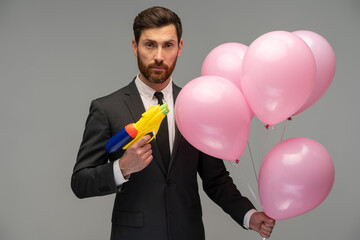 Serious businessman wearing formal suit holding toy gun and a lot of balloons with light smile, greeting on holiday and sharing present. Indoor studio shot isolated on grey background