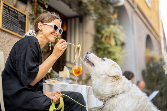 Woman eating pasta with her cute white dog at the restaurant outdoors. Concept of friendship with pets and having fun together