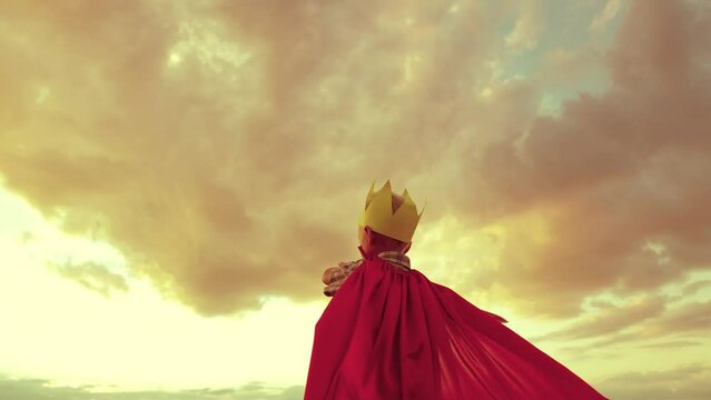 boy plays superhero against backdrop sunset sky. small child kid red cape with sword his hand. child dream kid. boy golden crown plays game. costume children's fantasy happy baby vacation holiday.