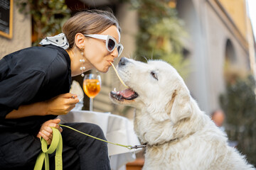 Woman eating pasta with her cute white dog at the restaurant outdoors. Concept of friendship with pets and having fun together