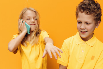funny, happy children, brother and sister are standing on a yellow studio background and the girl is talking on her phone. Horizontal photo with empty space for text insertion