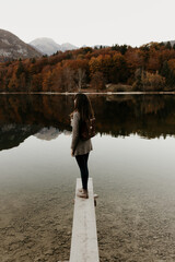 A woman is standi on a wooden plank by a lake in Slovenia. The water is cristal clear. The woman is wearing gray cardigan and a brown leather backpack. The mountains are reflecting on the water. Fall.