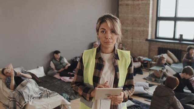 Medium portrait of young Biracial female volunteer wearing green reflective vest, holding tablet computer, standing in foreground of refugees in shelter at daytime, woman looking on camera