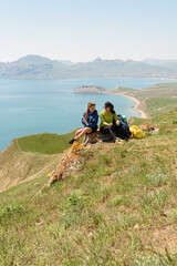 Two young beautiful woman travel with backpacks, sit on mountains view and drink tea smiling each other