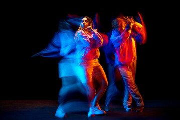 Fototapeta premium Dynamic portrait of stylish man and girl dancing hip-hop in sportive style clothes on dark background at dance hall in mixed neon light. Youth culture, breakdance, movement