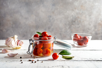 Pickled tomatoes in a glass jar with garlic, oil, pepper and bay leaves on white wooden table with grey background