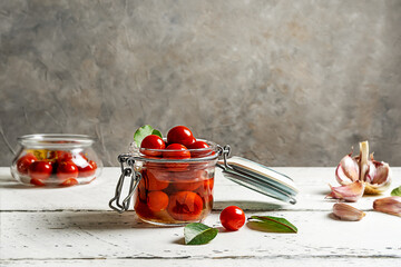 Pickled tomatoes in a glass jar with garlic, oil, pepper and bay leaves on white wooden table with grey background