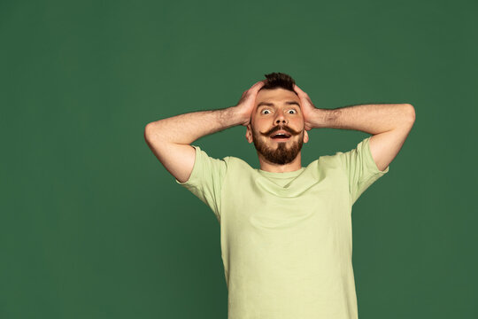 Portrait of young emotive man with moustache posing with shocked face isolated over green studio background