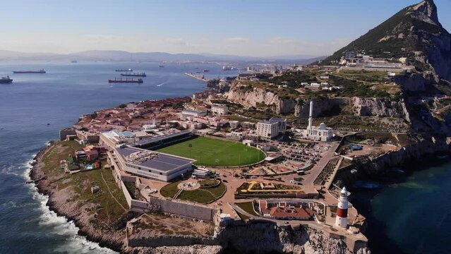 Green stadium of Gibraltar national rugby union team at Europa point with the recognizable lighthouse above the high cliffs and rocks where the waves crash on a sunny day. Backwards drone dolly shot