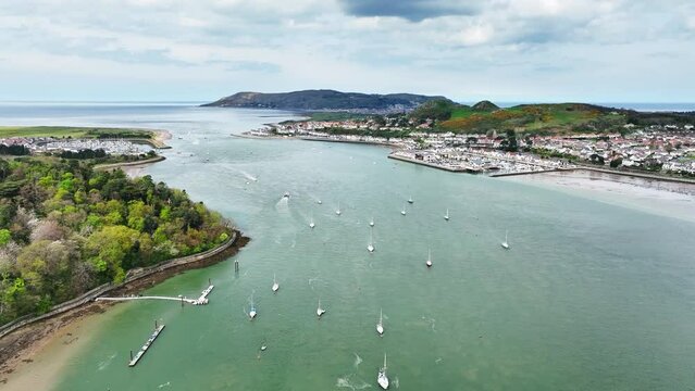 Several sailboats anchor in the fast flowing Conwy River with the coastal town of Deganwy amongst the green hills of Welsh nature on a cloudy day. High drone dolly shot