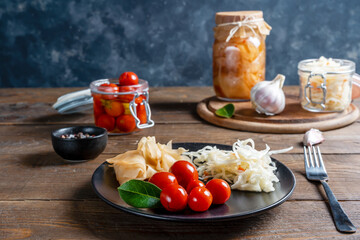 Pickled tomatoes, kimchi, cabbage in jar on cutting board and plate on wooden table with grey background