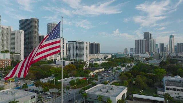 Fly around American national flag waving on pole above city. High rise office or apartment buildings in background. Miami, USA
