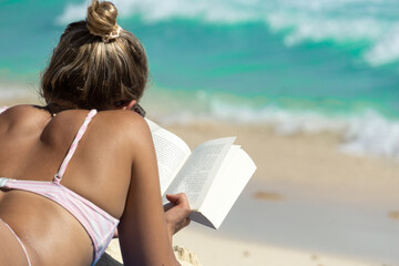 Reading on the beach. A tanned girl in a bikini lying on the sand reads a book, against the backdrop of blurry waves.