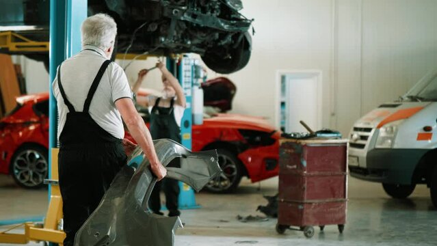 Old mechanic brings a new bumper to check if it fits on the damaged car in the workshop.