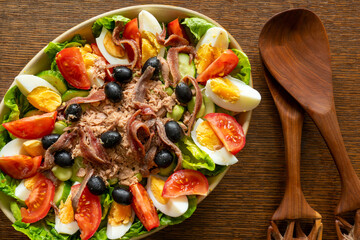 Homemade French salad, salad nicoise, in a porcelain dish on a wooden worktop with serving spoons. Top view.
