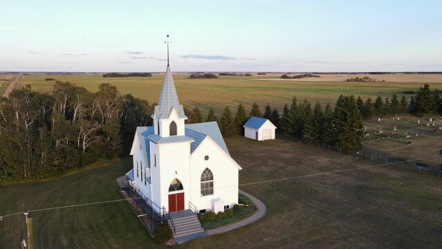 An ancient white church with a blue roof standing isolated in Canada's rural countryside. Wide angle aerial orbit