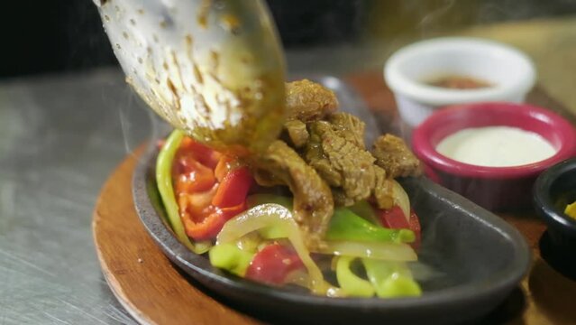 Chef is putting cooked meat with spoon on a black ceramic lava Mexican Fajita guvec plate in restaurant kitchen. slow motion,4K. Mexican Meat Food Fajita on a Plate.