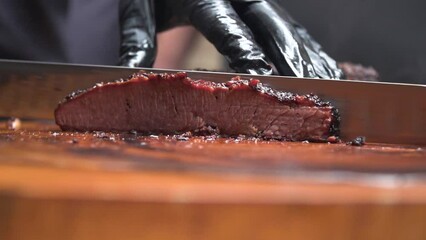 Slow motion: a professional chef cutting a tasty steak after cooking it, slicing the meat with a sharp knife on a wooden chopping board, wearing black gloves. Closeup side shot.