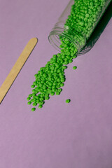 Beautiful green wax granules for depilation are poured from a transparent jar on a light purple background, a wooden spatula. Epilation, depilation, removal of unwanted hair. View from above.