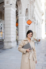 Fototapeta premium happy woman in trench coat walking with laptop on street of vienna.