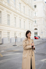 Fototapeta premium cheerful woman in beige trench coat walking with laptop on street in vienna.