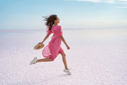Running carefree woman on inspiring landscape of salt flats on pink lake. Brunette model in pink adorable flowing dress