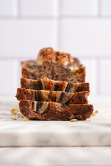 Slices of Chocolate banana bread with walnuts on a marble board and ingredients on a grey neutral background