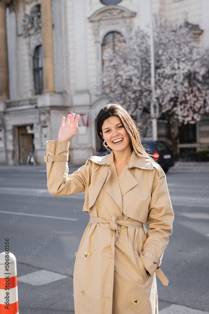 Wall mural happy woman in elegant coat smiling and waving hand on street in vienna.