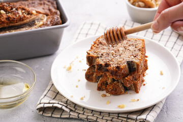 pouring honey onto Slices of Chocolate banana bread with walnuts on a checkered kitchen napkin and ingredients on a grey neutral background