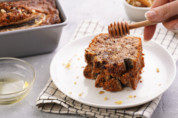 pouring honey onto Slices of Chocolate banana bread with walnuts on a checkered kitchen napkin and ingredients on a grey neutral background
