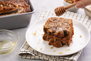 pouring honey onto Slices of Chocolate banana bread with walnuts on a checkered kitchen napkin and ingredients on a grey neutral background