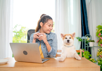 The dog sit and put its leg on table also stay beside pretty Asian woman using tablet and also look at her pet with happiness. The dog also look at camera.