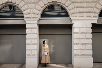 full length of happy woman in beige trench coat standing with laptop near arch.