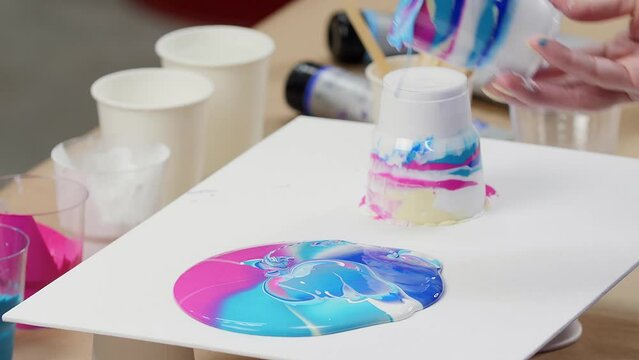 Woman creates modern art, pouring paint from glasses on a sheet of paper in an original way. Acrylic paint circles spread. Spilled paint. Drawing concept.