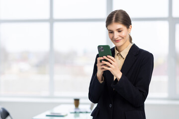 Obraz premium Smiling businesswoman using phone in office. Small business entrepreneur looking at her mobile phone and smiling.